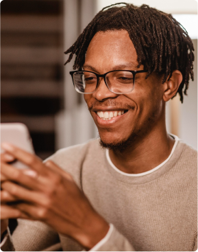 A picture of a young black adult male with dreads and eyeglass, he looks into his mobile phone smiling.